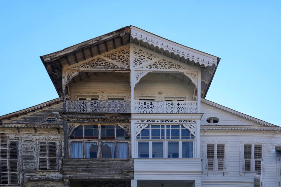 A two-story house seen during a house relocation process, with the bottom half displaying weathered, dark wood siding and old, closed window shutters, while the upper half has recently been painted wh