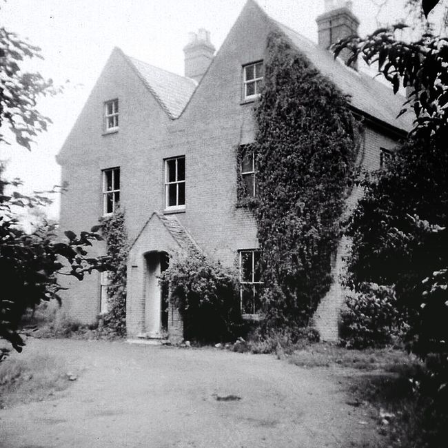 A two-story house seen during a house relocation process, with the bottom half displaying weathered, dark wood siding and old, closed window shutters, while the upper half has recently been painted wh