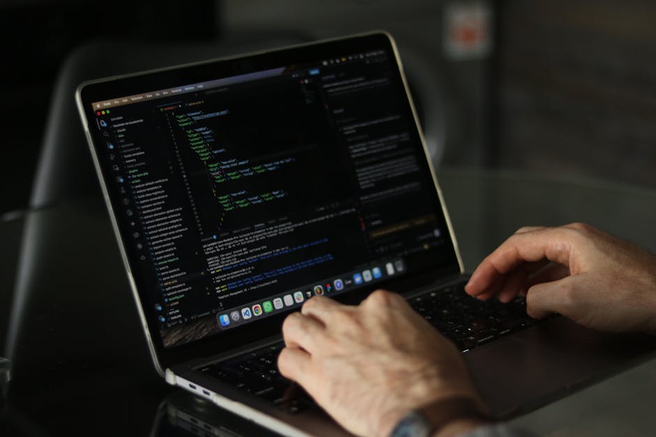 Close-up view of a person's hand typing on a laptop keyboard displaying code or scripting language, resting on a wooden desk. In the background, part of the laptop screen shows lines of colorful code,