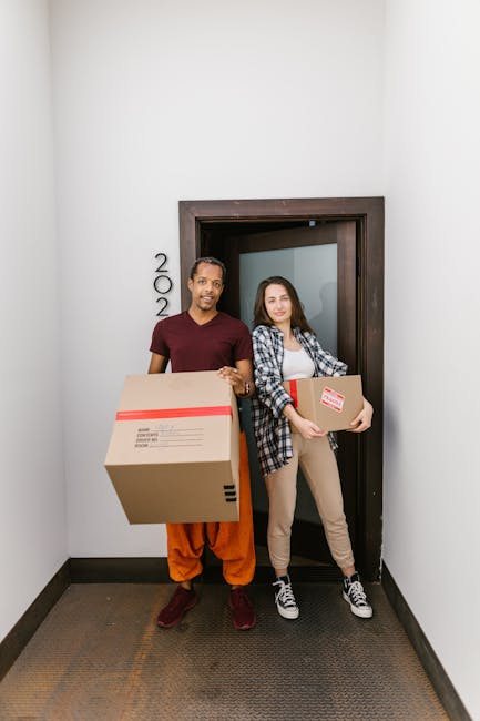 A group of three diverse young adults, two women and one man, are outside a building during daylight, smiling and taking a selfie with a smartphone. They are holding multiple cardboard boxes sealed wi