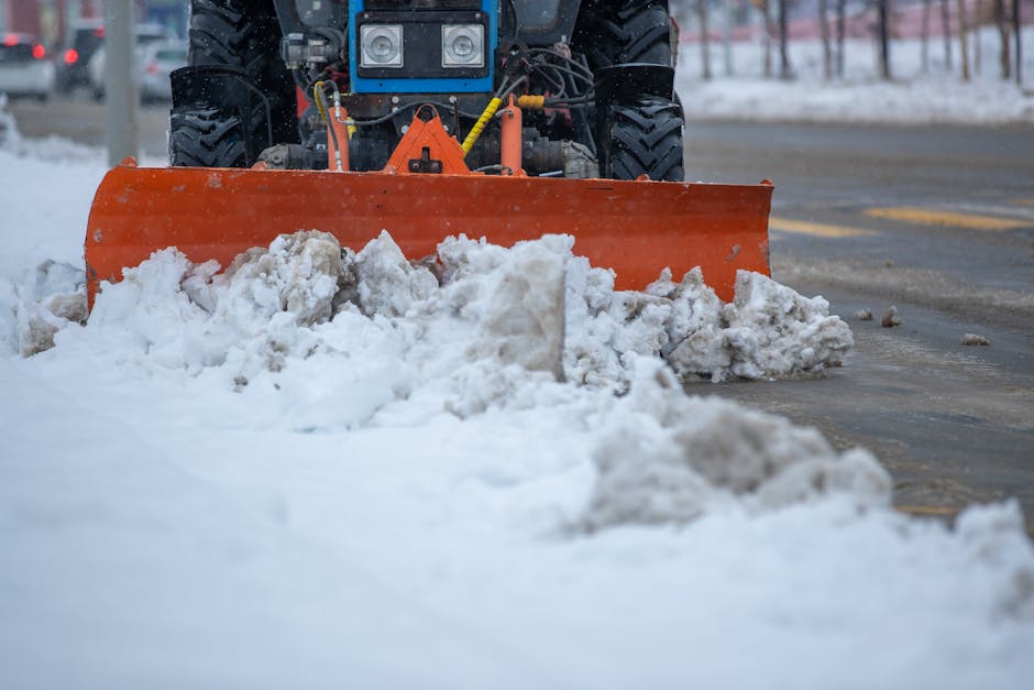 A snow plough with a bright orange blade clearing snow from a paved street during daytime, with the vehicle's front wheels visible and the blade pushing accumulated snow aside. The snow is partially m